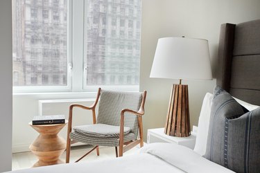 Modern bedroom with gray upholstered headboard, wood lamp, nightstand, armchair, side table.